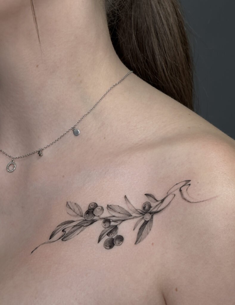 Delikatny tatuaż fine line przedstawiający gałązkę oliwną z liśćmi i owocami, wykonany cienką linią na obojczyku. Subtelny, minimalistyczny tatuaż botaniczny w czerni, idealny dla osób ceniących eleganckie i kobiece wzory.