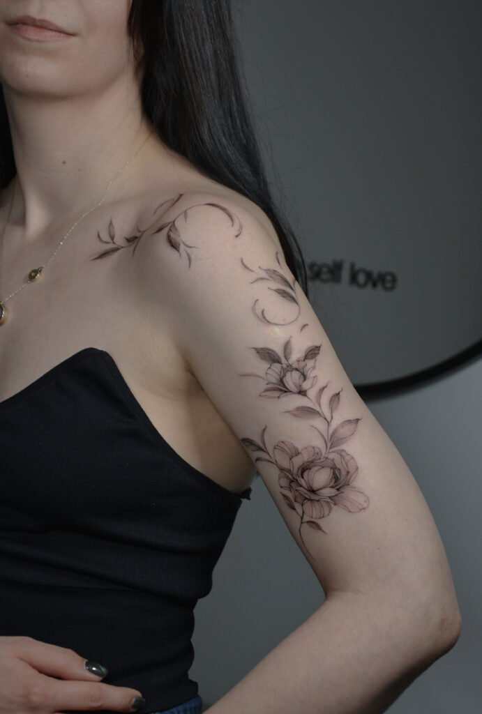 Elegancki tatuaż kwiatowy w stylu fine line, wykonany cienką linią z delikatnym cieniowaniem, obejmujący ramię i górną część ręki. Subtelny tatuaż botaniczny w czerni, podkreślający kobiecą formę i naturalny ruch linii.